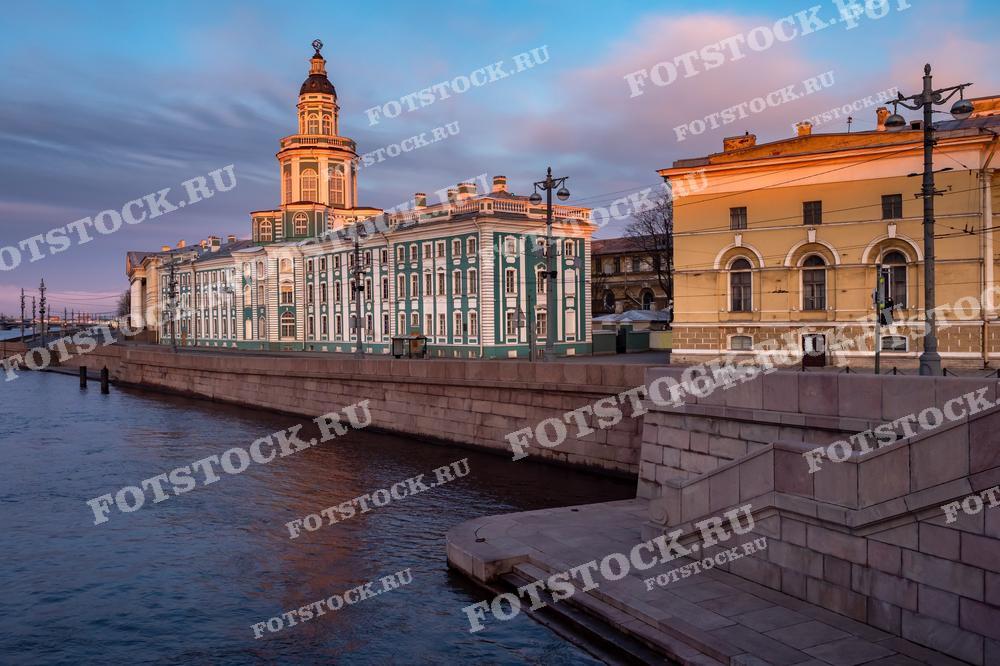 На фотографии изображено здание Кунсткамеры в Санкт-Петербурге во время восхода или заката солнца, что видно по тёплым цветам неба и отражению солнечного света на фасаде здания и поверхности реки. Мягкий свет подчеркивает архитектурные детали здания и создаёт тихую, умиротворённую атмосферу. Фотография может быть использована для туристических путеводителей, исторических книг, материалов по искусствоведению и культуре, а также в качестве декоративного элемента в интерьерах.
